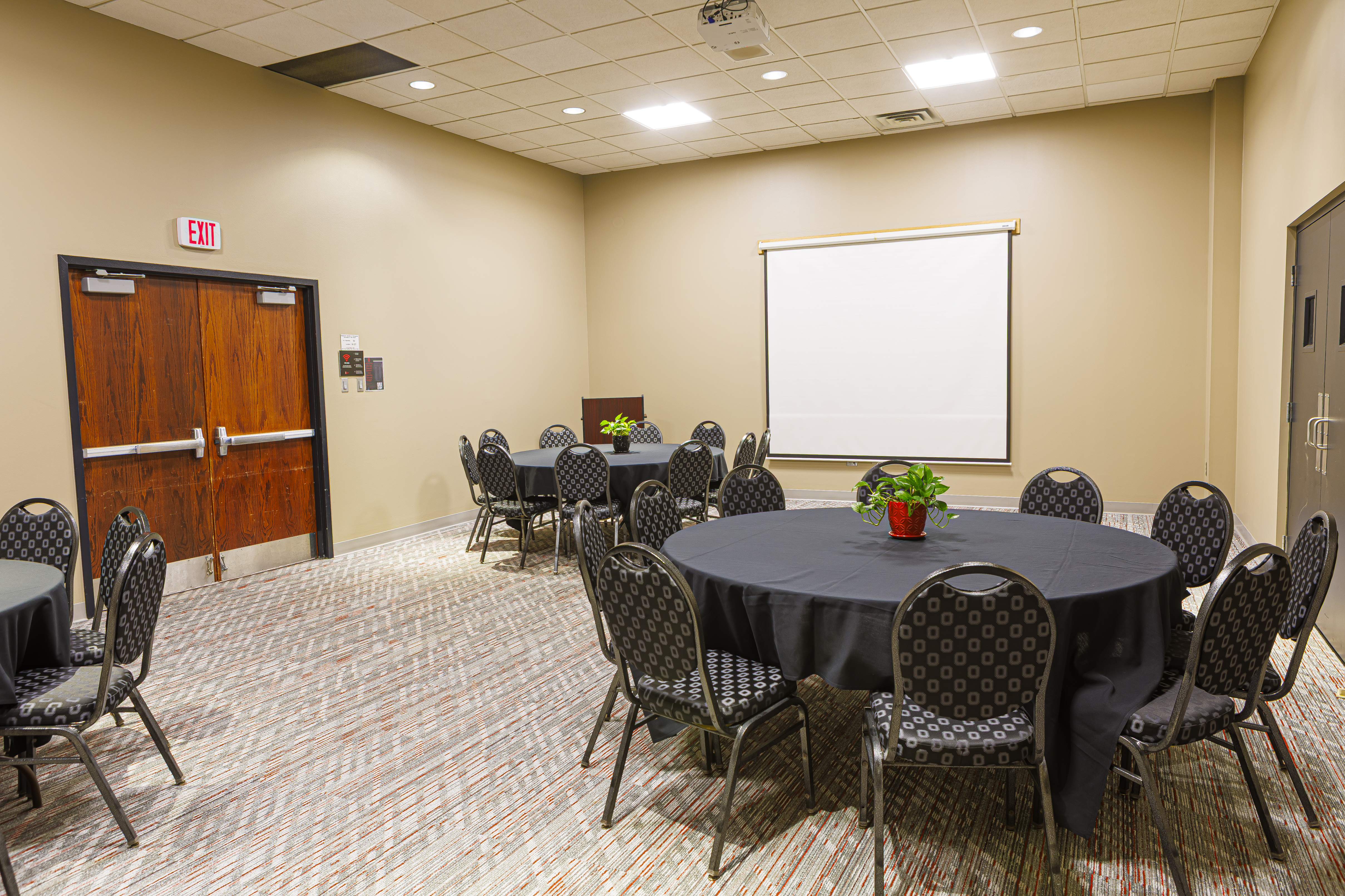 Meeting room with round tables, black linens, a projector screen, and a dark wood door near the left wall.