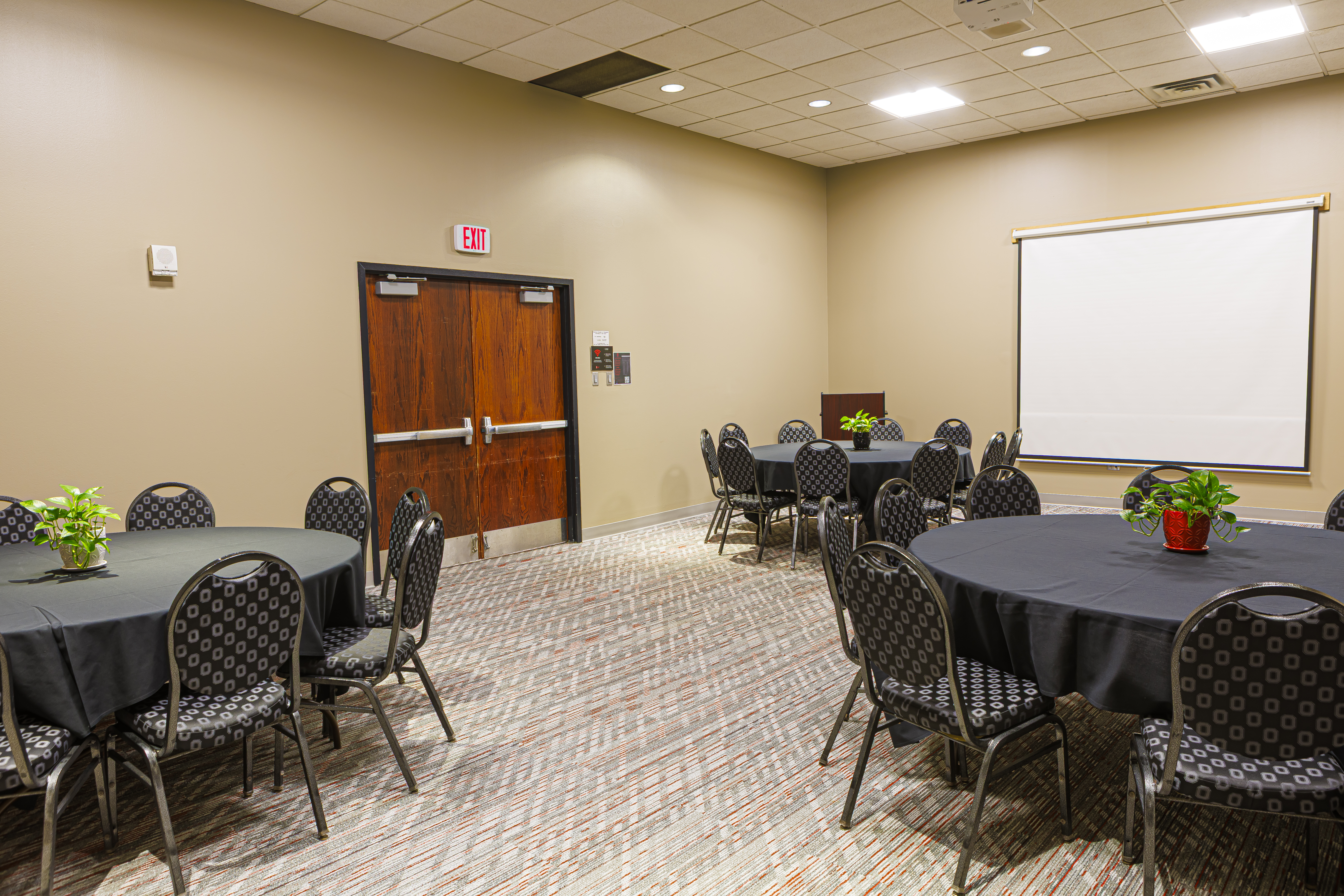 Meeting room with round tables, black linens, a projector screen, and a dark wood exit door on a tan wall.