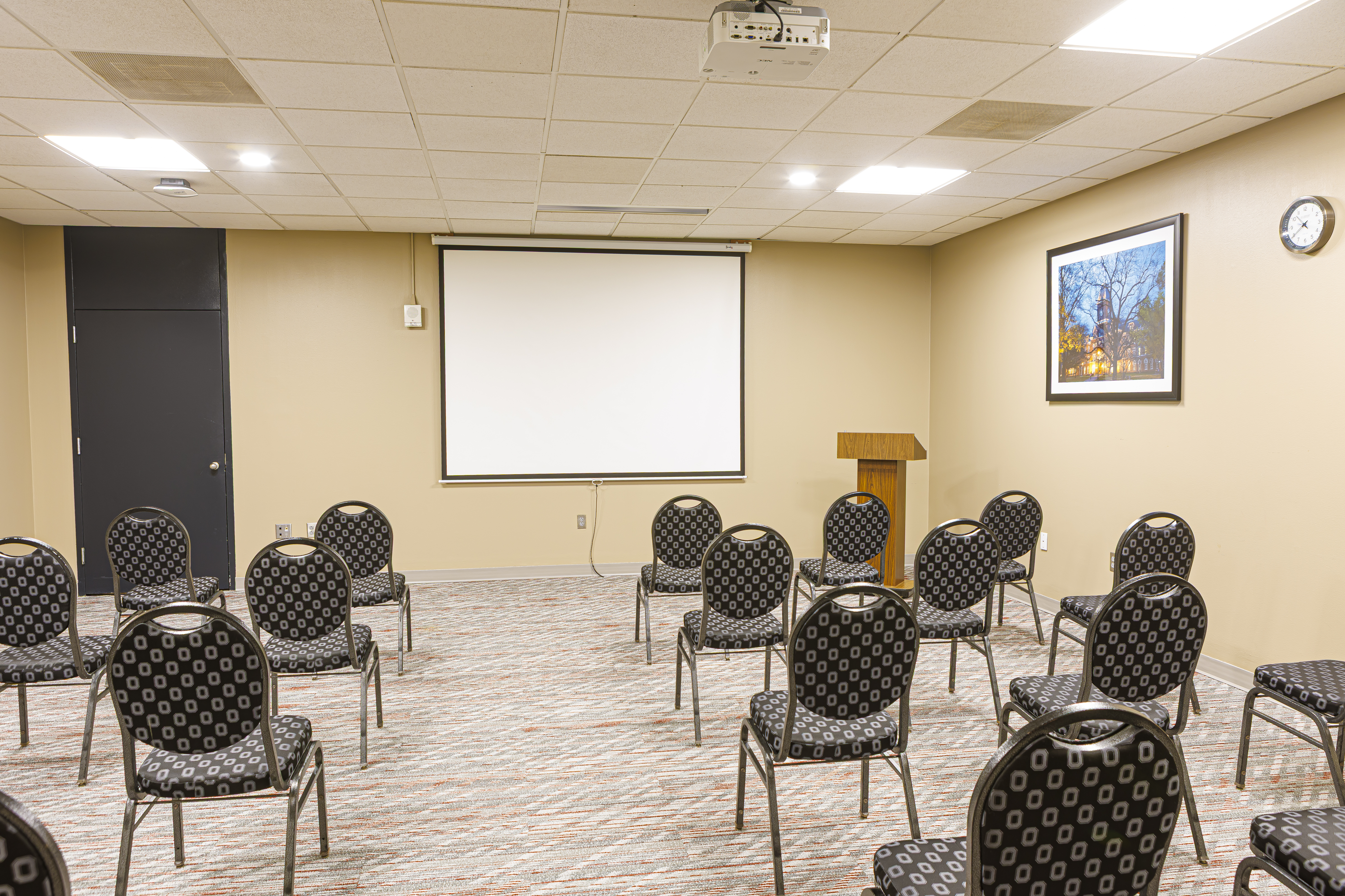 Small meeting room with patterned chairs facing a projector screen, a wooden podium, and a framed photo on a tan wall.