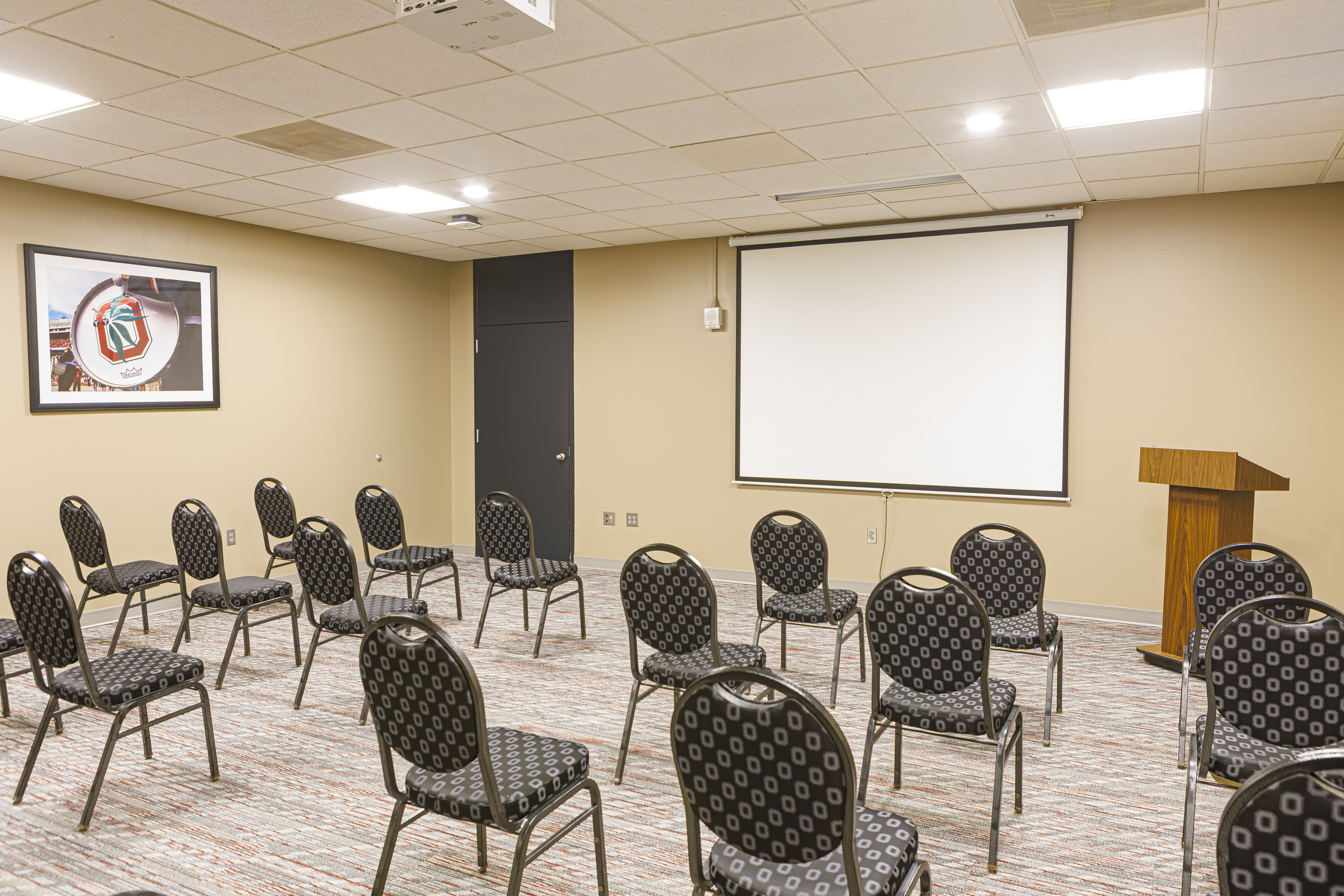 Small meeting room with scattered chairs facing a projector screen and podium. An OSU-themed photo hangs on the left wall.