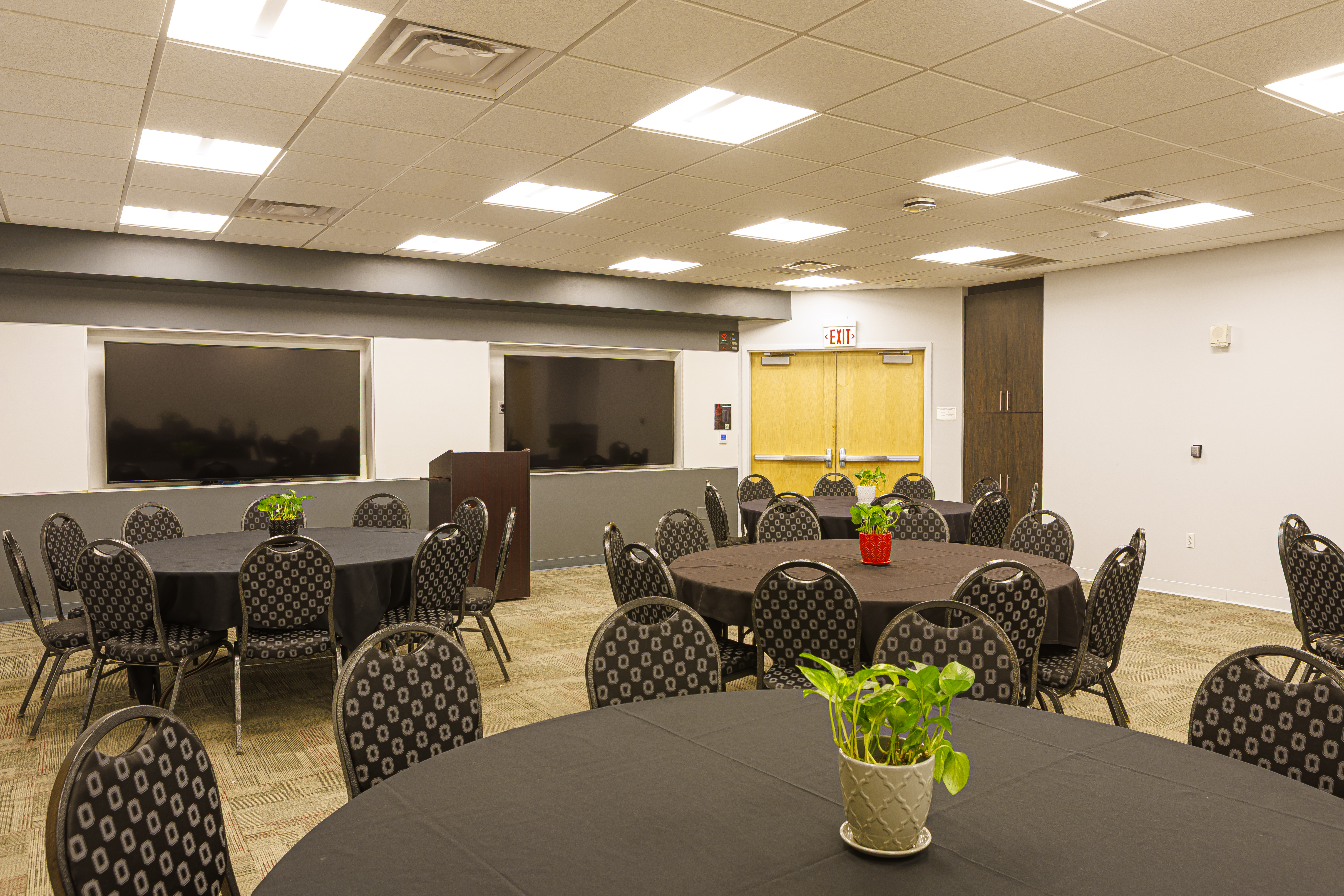 A meeting room with several round tables draped in black linens and patterned chairs. Two wall-mounted TVs face the room.