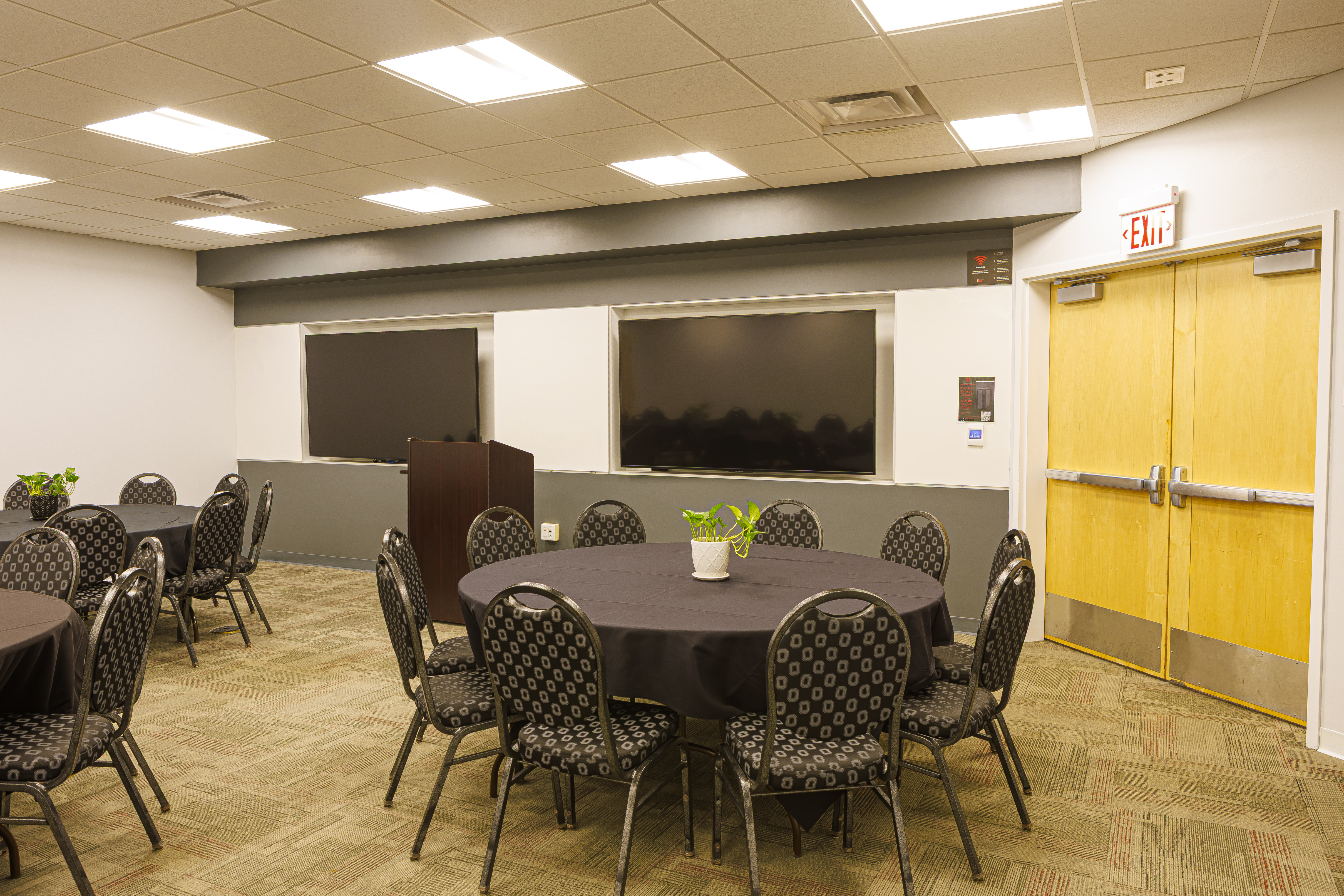 A meeting room with round tables, black tablecloths, and patterned seating. A podium stands between two TVs.