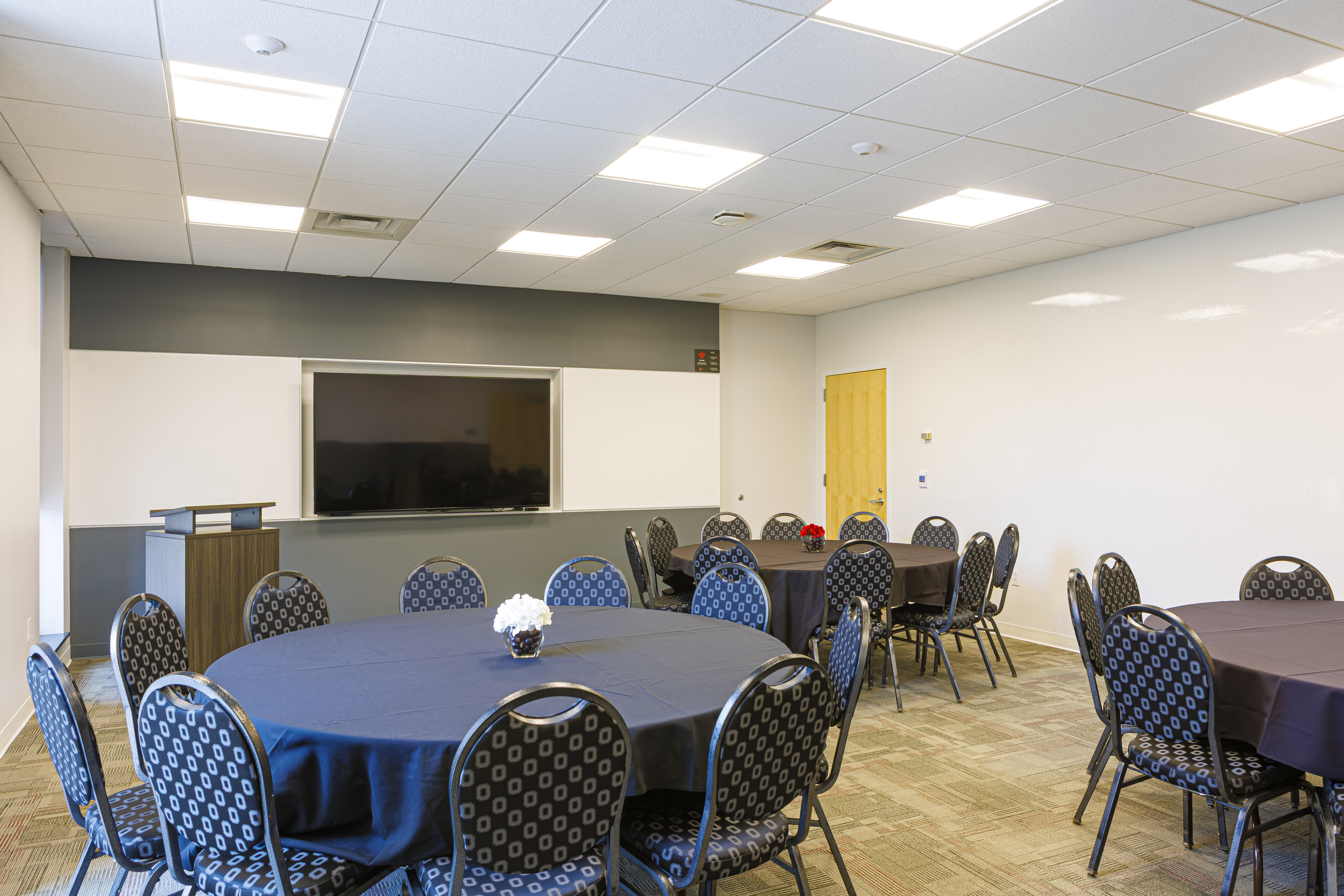 A bright room with three round tables, black linens, and black-patterned chairs. A single large TV hangs on the wall.