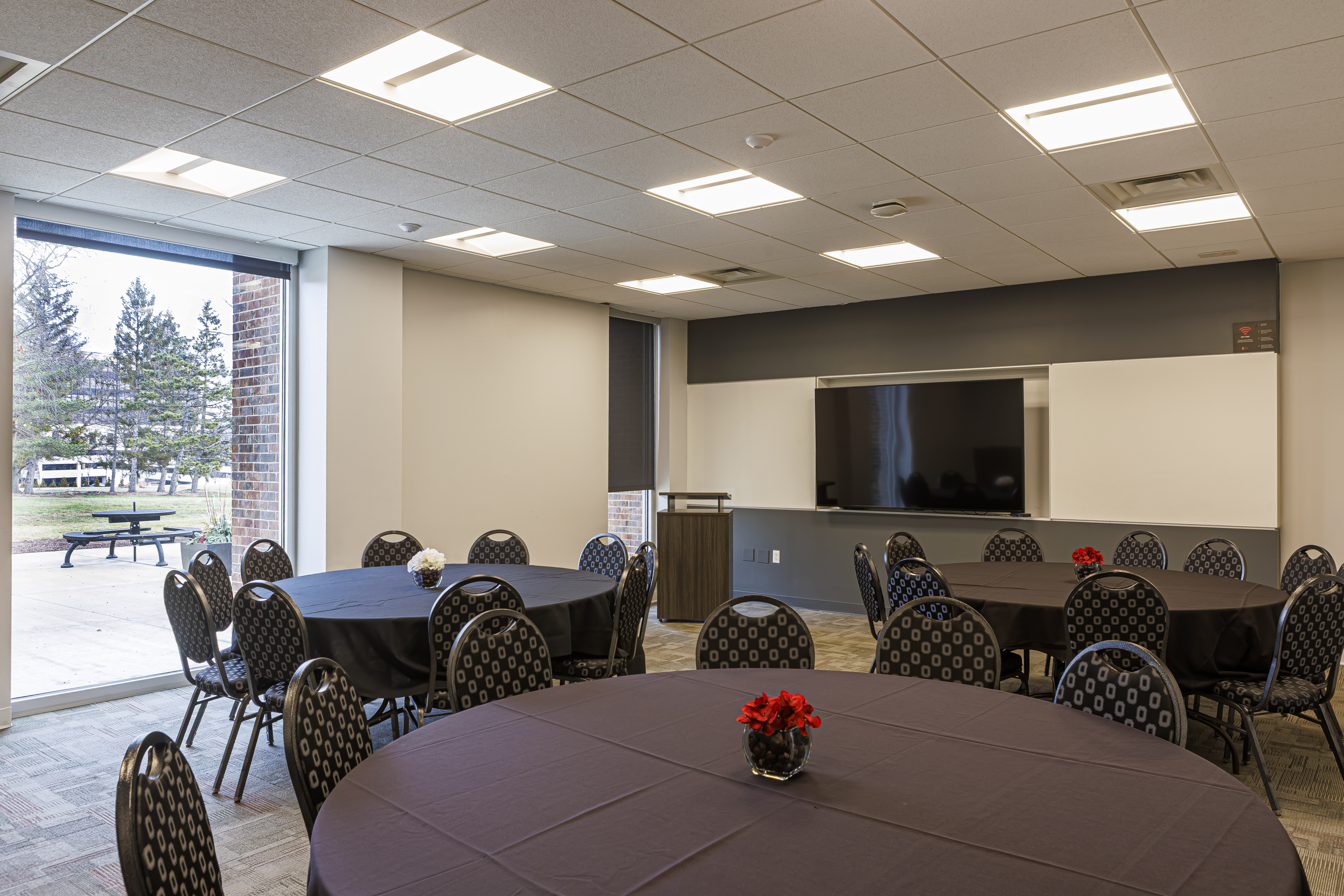 Meeting room with three round tables and black linens. A large TV and wood podium stand near a large window.