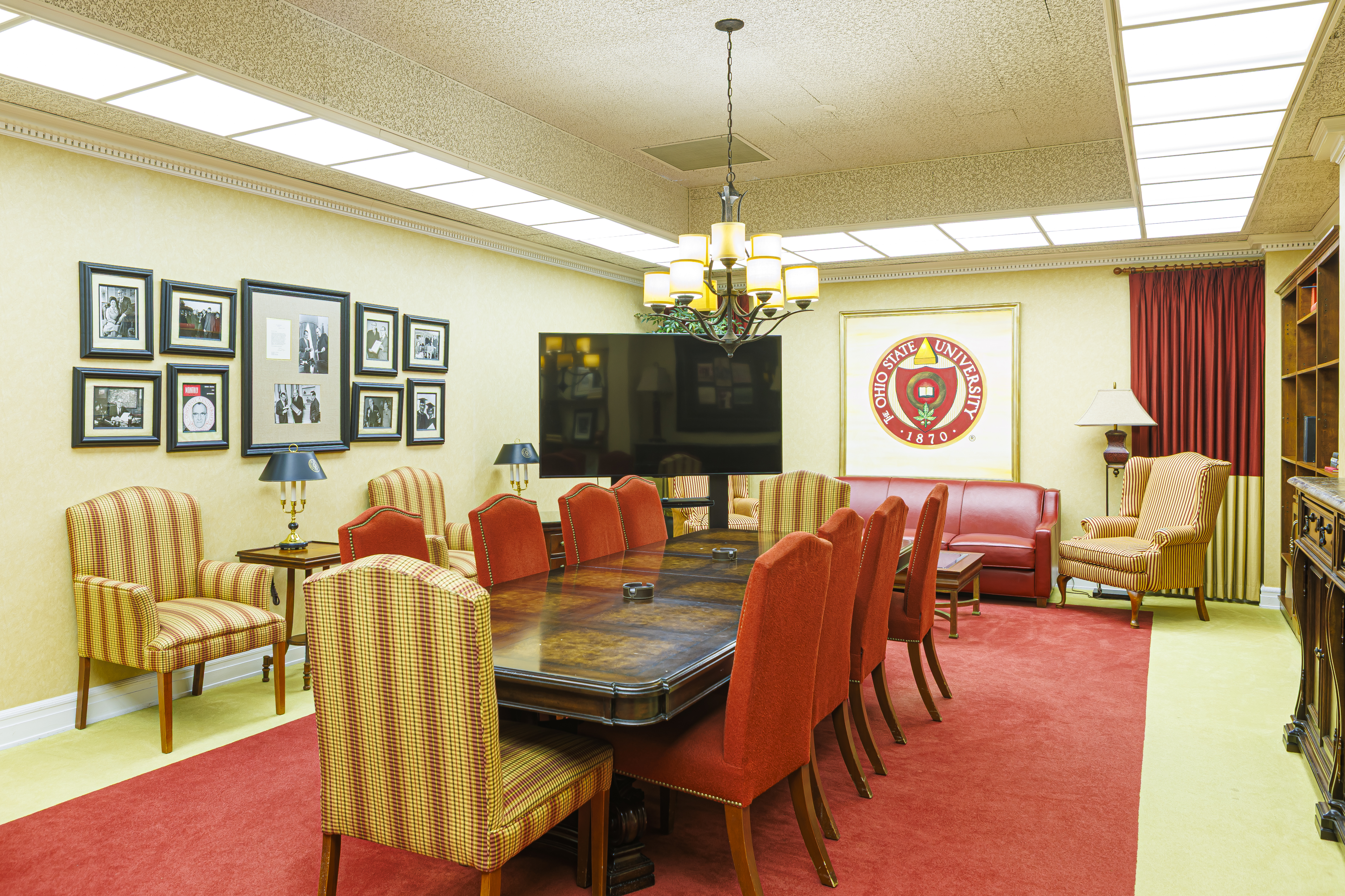 Elegant boardroom with a long wood table, red chairs, a chandelier, and a large university seal on the back wall.