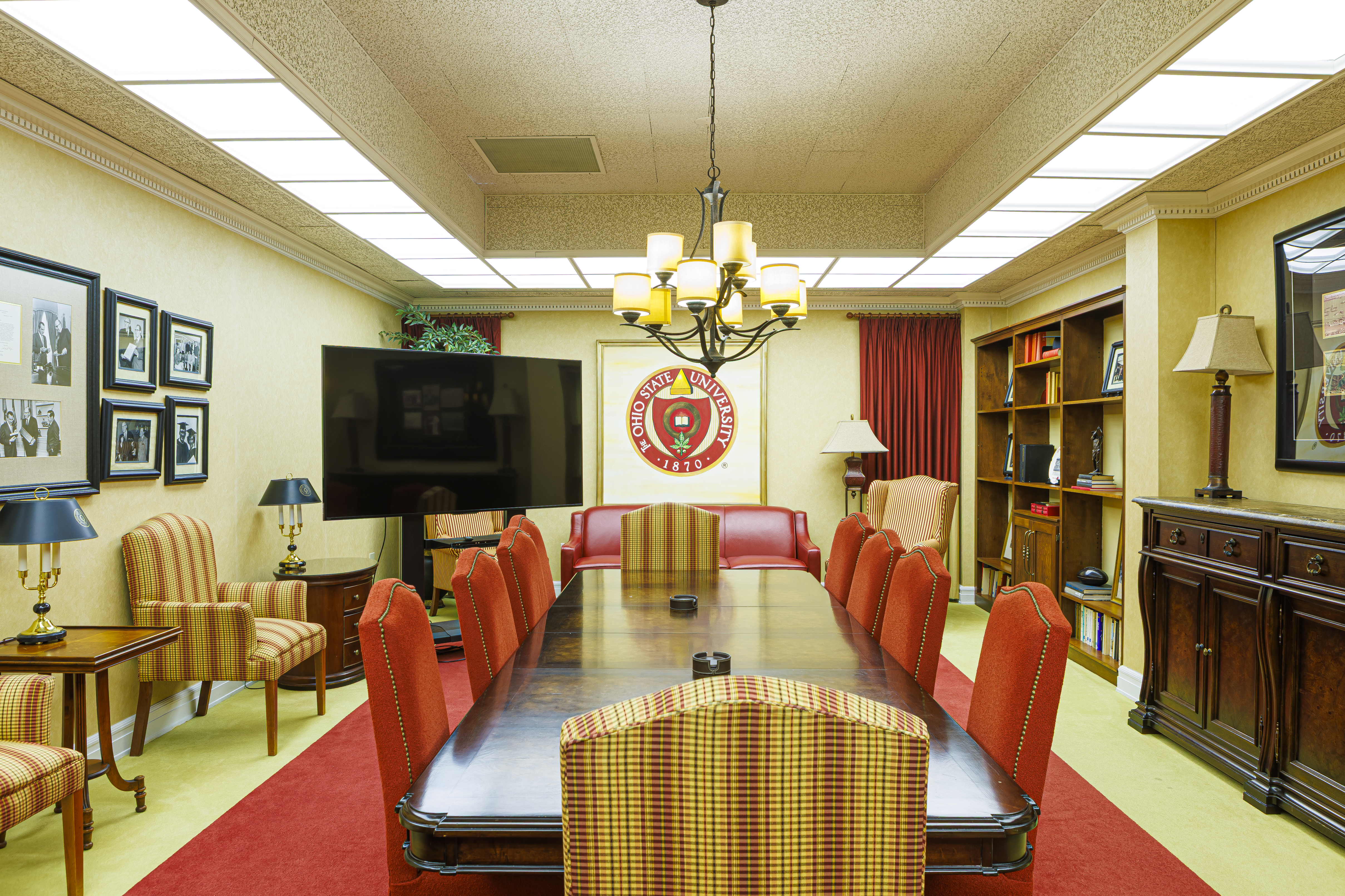 Boardroom with a long wood table, red chairs, and a chandelier. A large university seal is centered on the back wall.