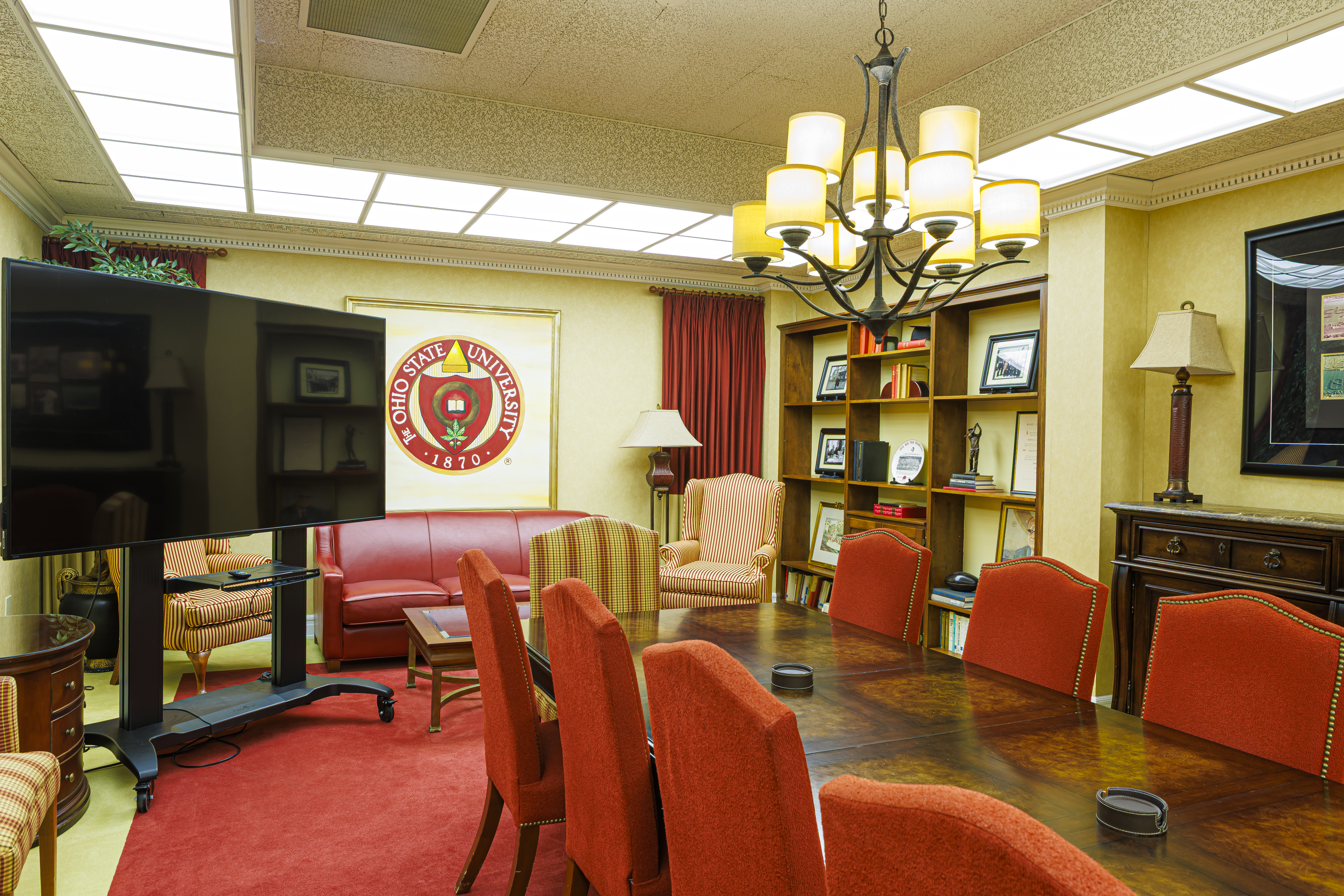 Close-up of a boardroom table and red chairs with a large TV, bookshelf, and university seal in the background.